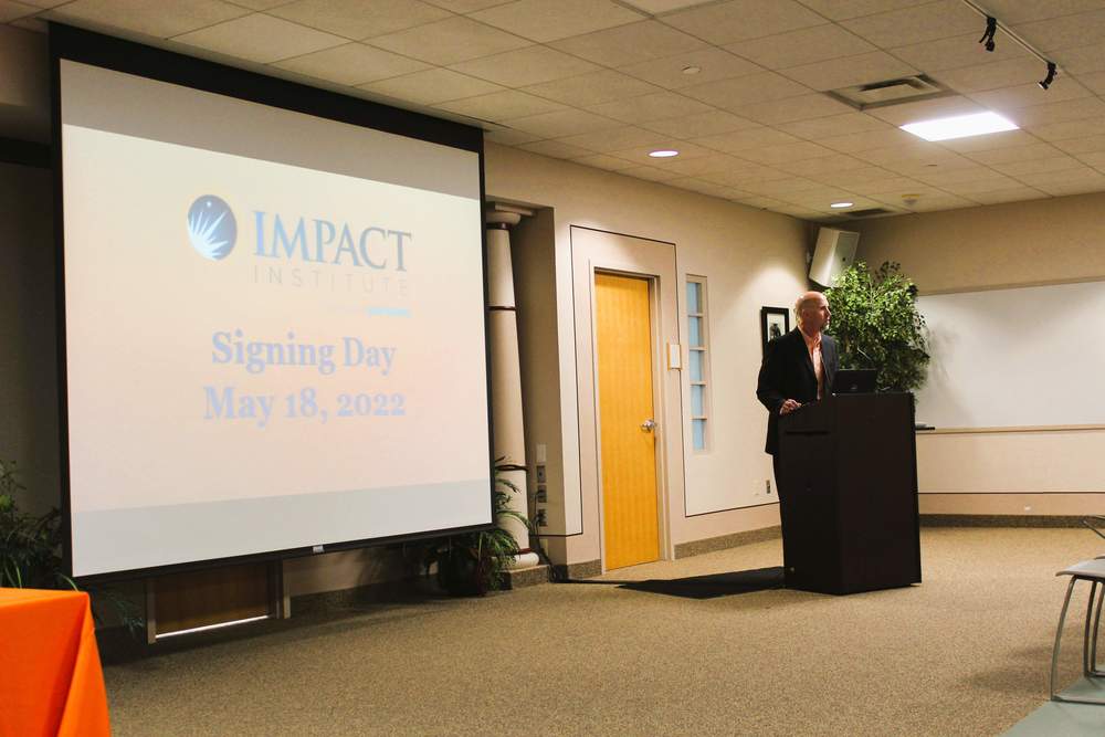 Impact signing day ceremony on projector screen with speaker at podium in front of screen