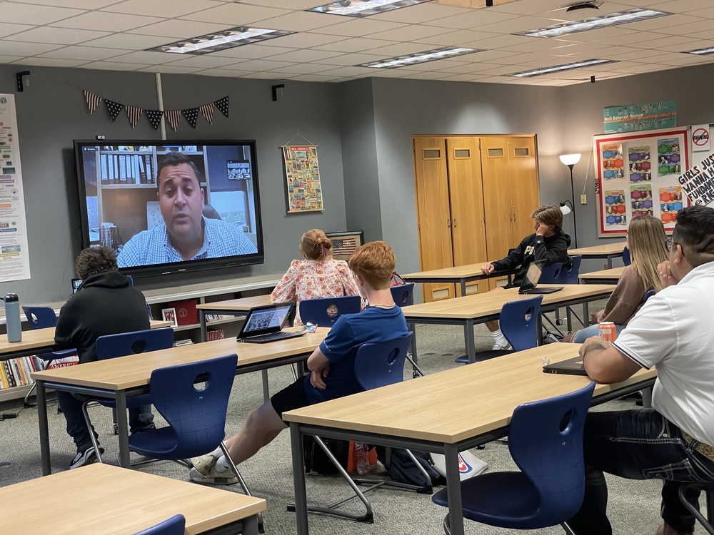 nine high school students watching a person speak on a large screen